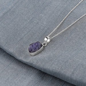 Raw blue tanzanite crystal pendant placed on fabric background showcasing natural rough cut and elegant bezel setting