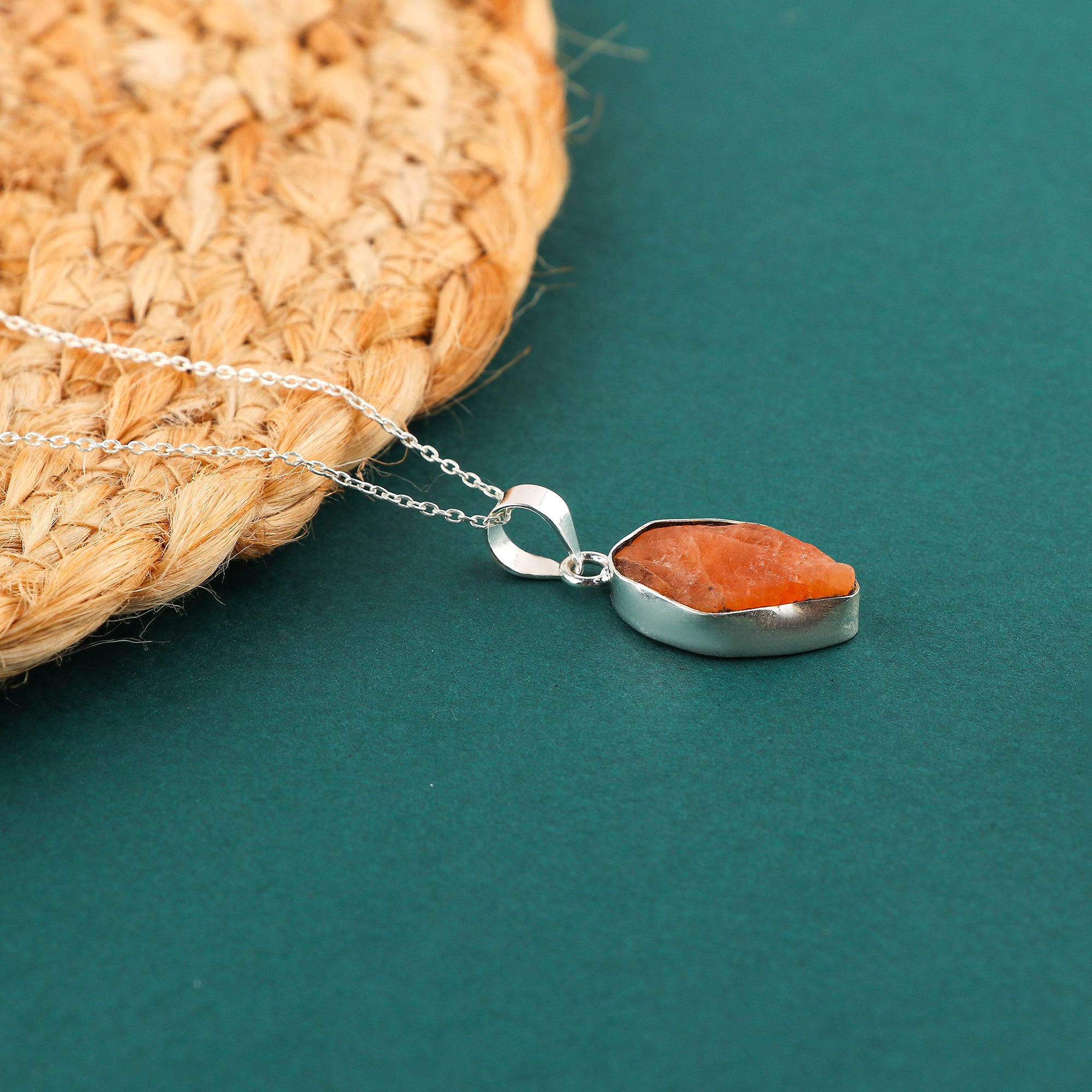 Natural orange triplite raw stone pendant displayed on a woven mat with silver chain highlighting its rough crystal texture