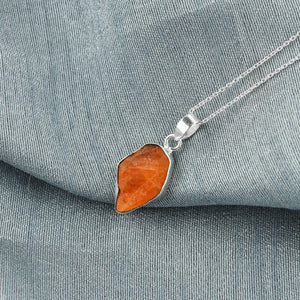 Triplite raw crystal pendant placed on grey fabric showing vibrant orange color, bezel setting and silver-plated chain details