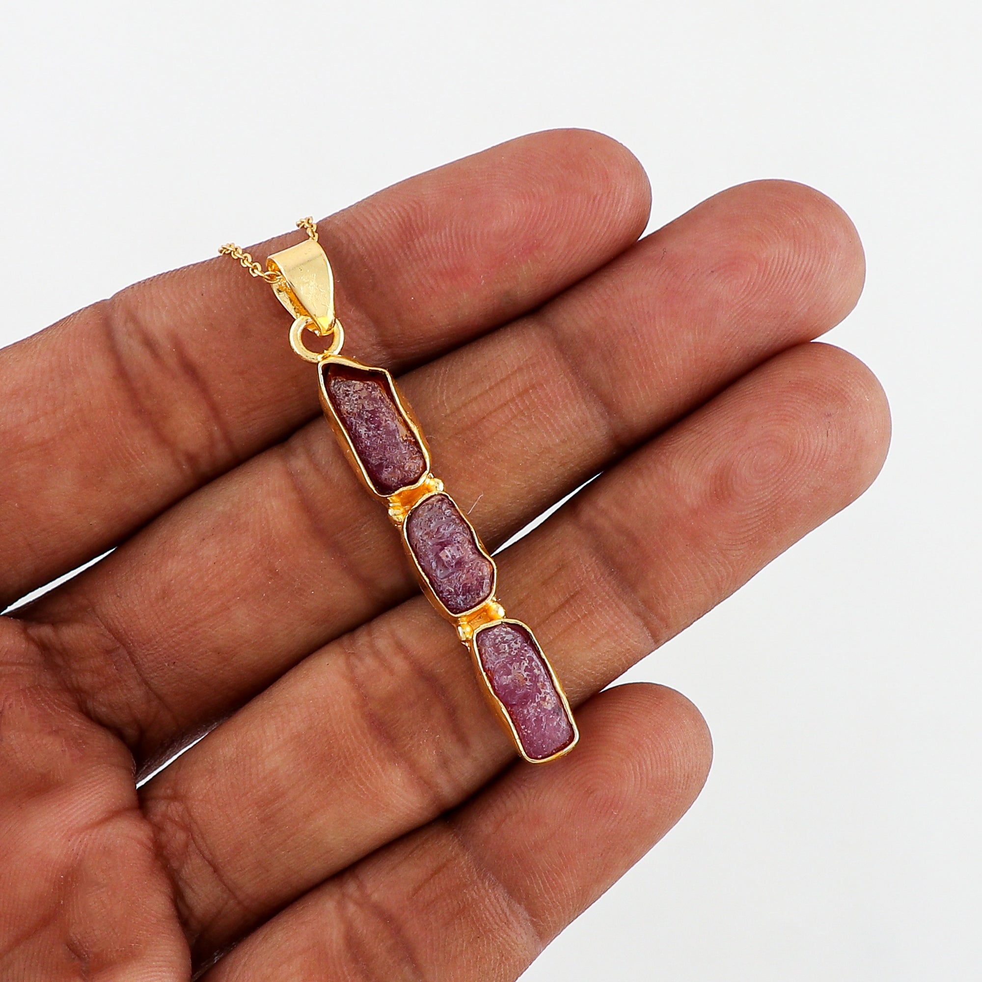 Natural pink ruby raw stone pendant held in hand showing irregular shape and gold-plated bezel on fine chain